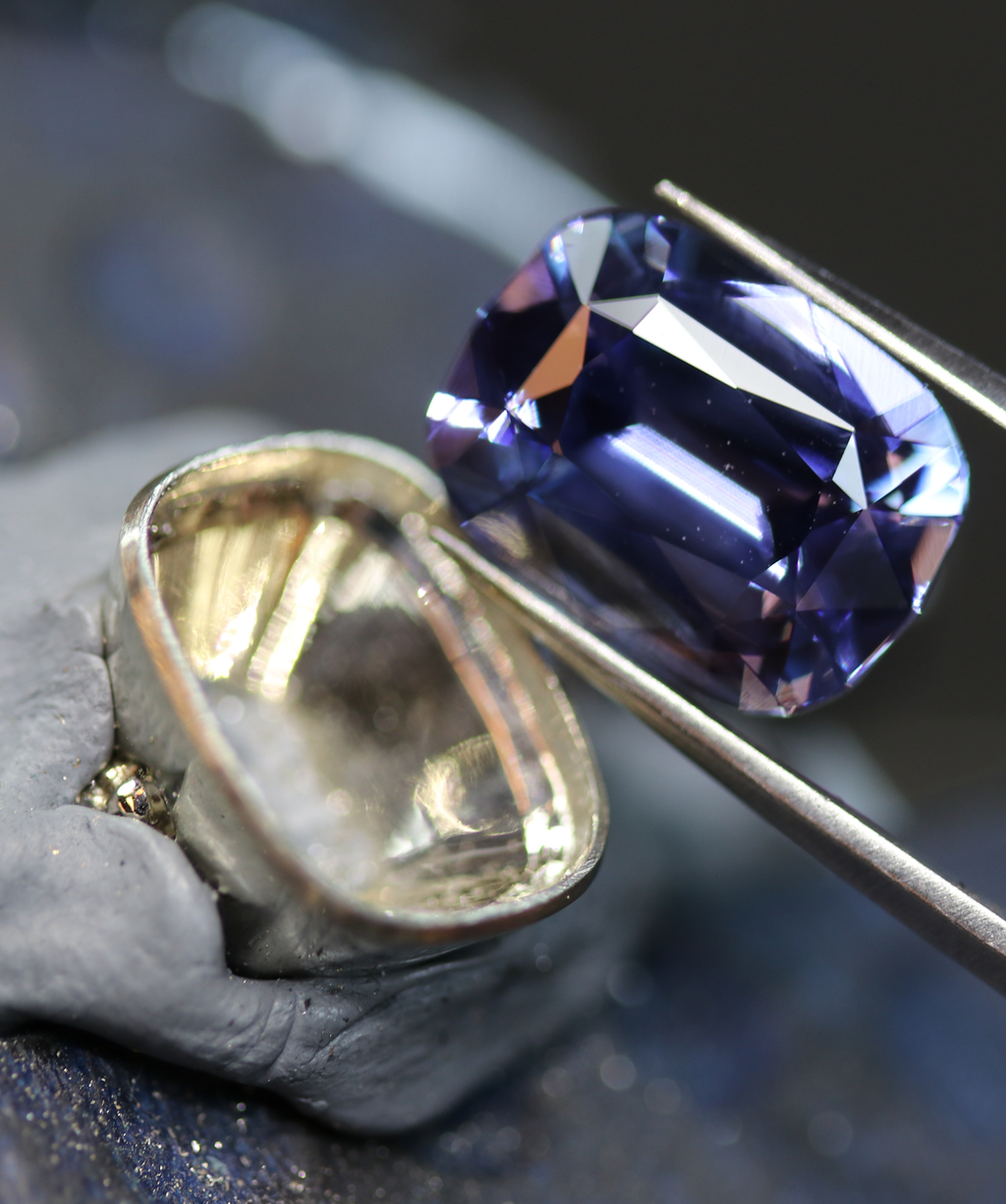 Photo shows a behind the scenes look at a jeweler working in close detail setting a stone in a bezel set engagement ring.