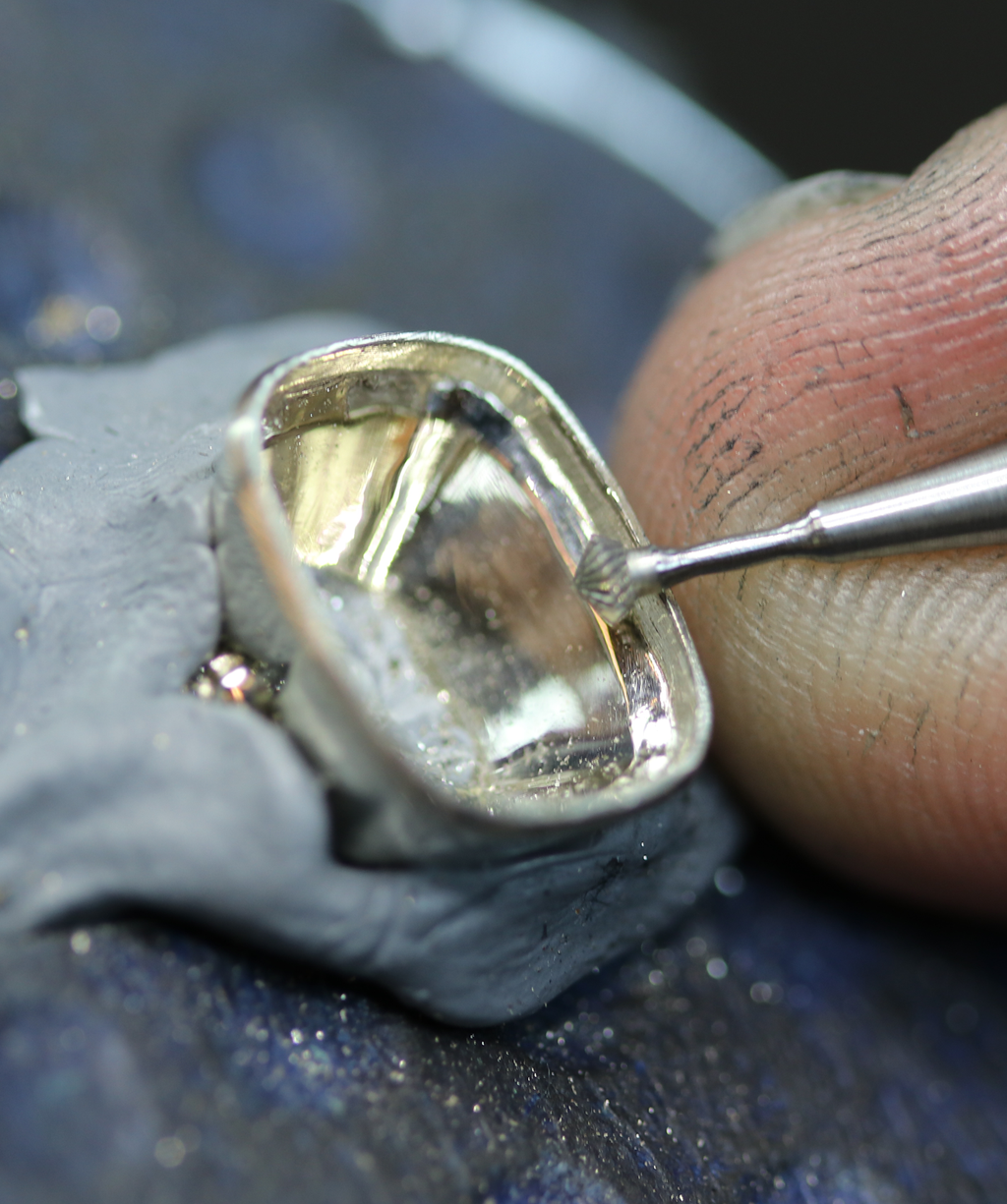 Photo shows a behind the scenes look at a jeweler working in close detail on the setting of a bezel set engagement ring.