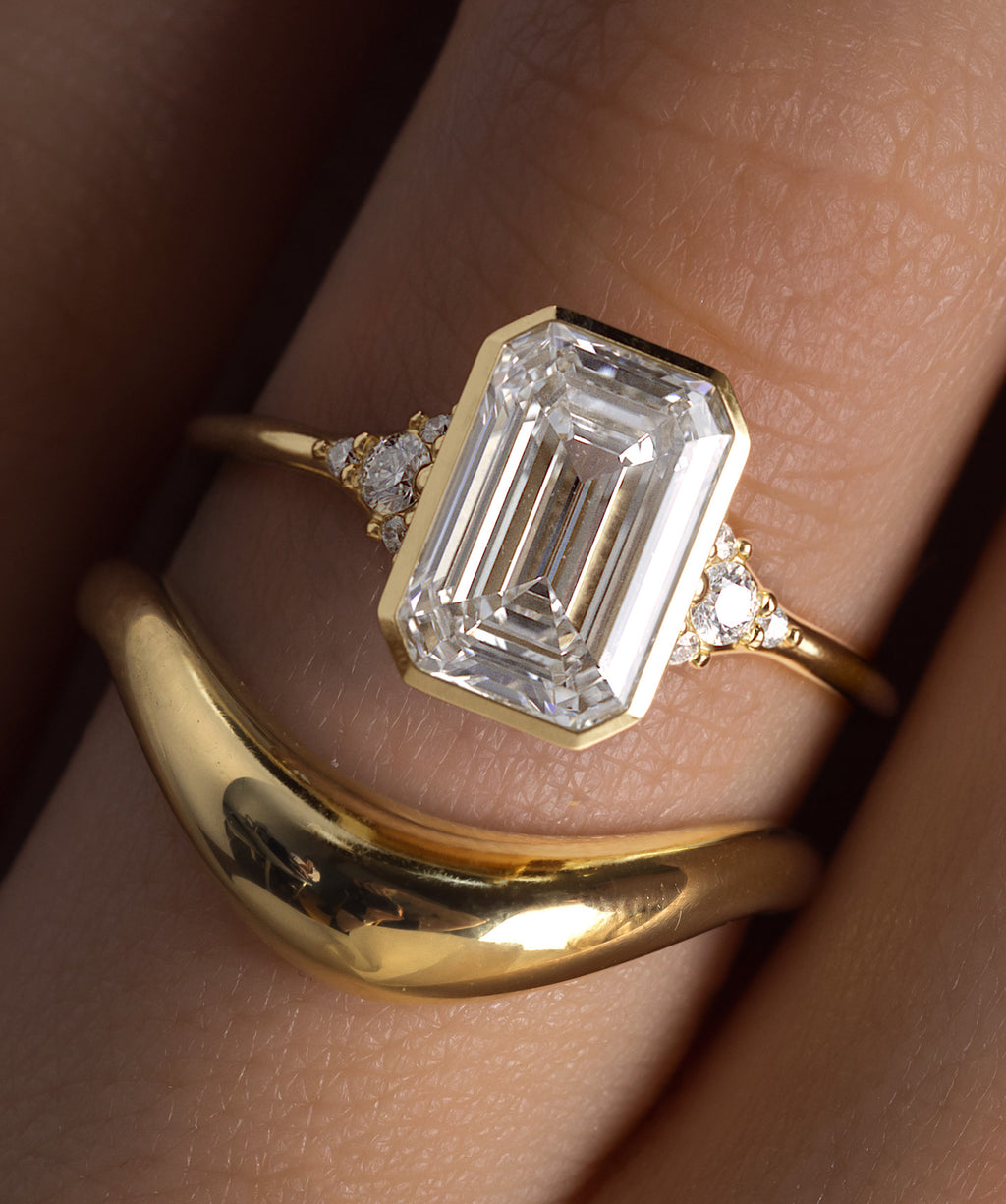 Photo features a model wearing an emerald cut Lady's Slipper Bezel Set Engagement Ring paired with an Amplify Band.