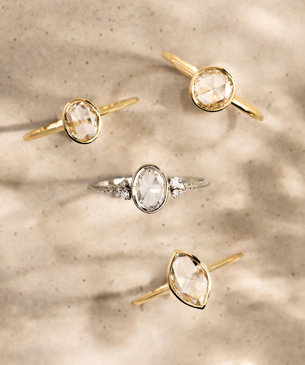 Photo features a variety of Melanie Casey Bezel Engagement Rings in Rose Cut from an above bird's eye view against a shadowy tan, speckled background.