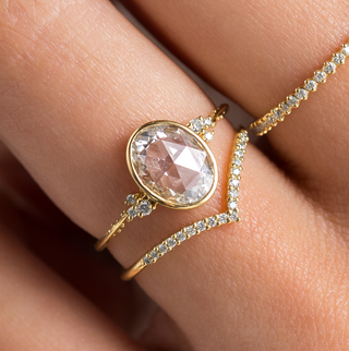 Photo features someone wearing the Bezel Engagement Pensive Ring in Oval and Rose Cut on her hand paired with a Chevron Wisp Band underneath and a Petite Many Moons Band on the next finger.